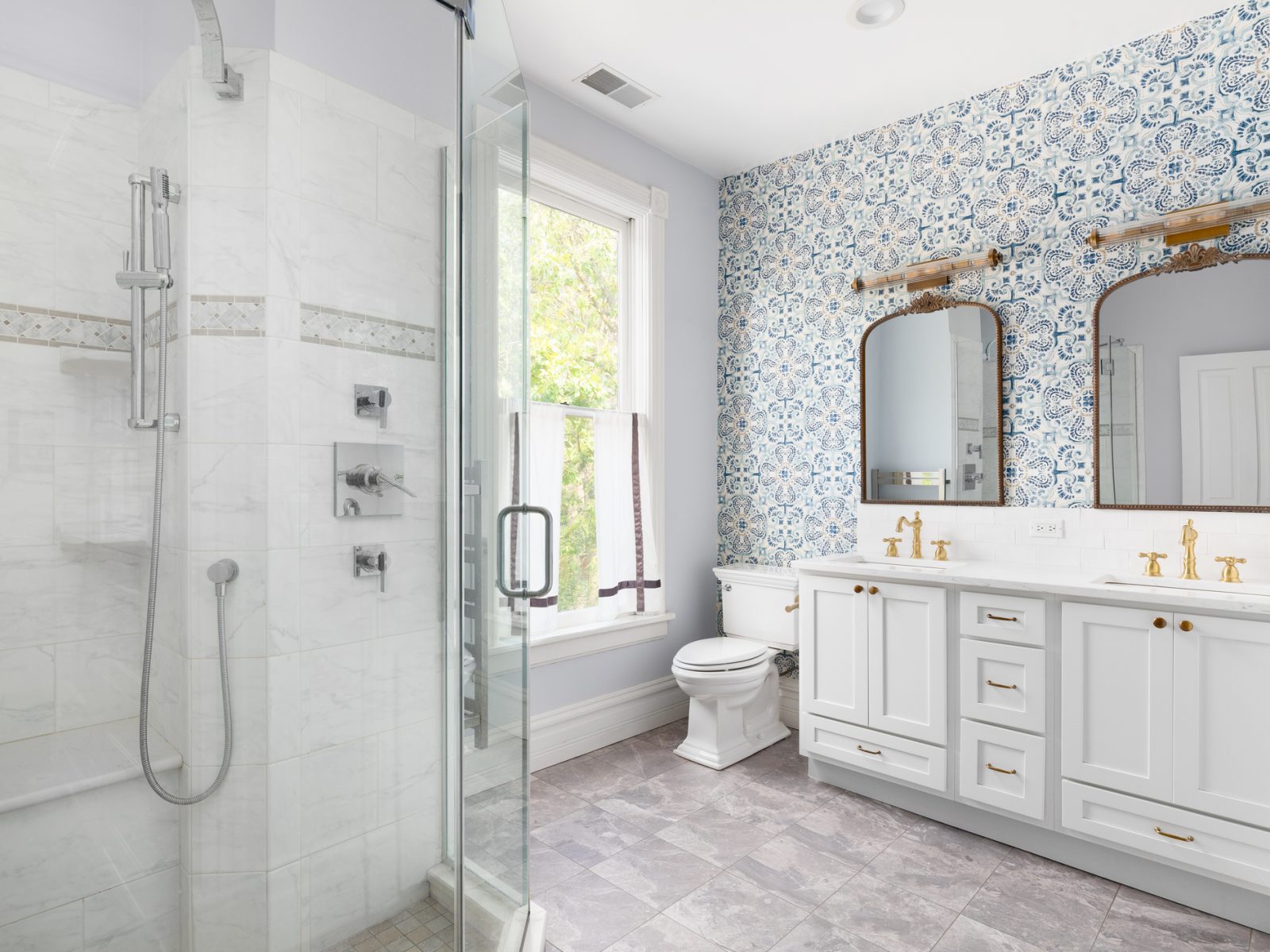 A cozy bathroom with a patterened blue wallpaper, grey cabinet, gold faucet and mirrors, and a tiled walk-in shower.